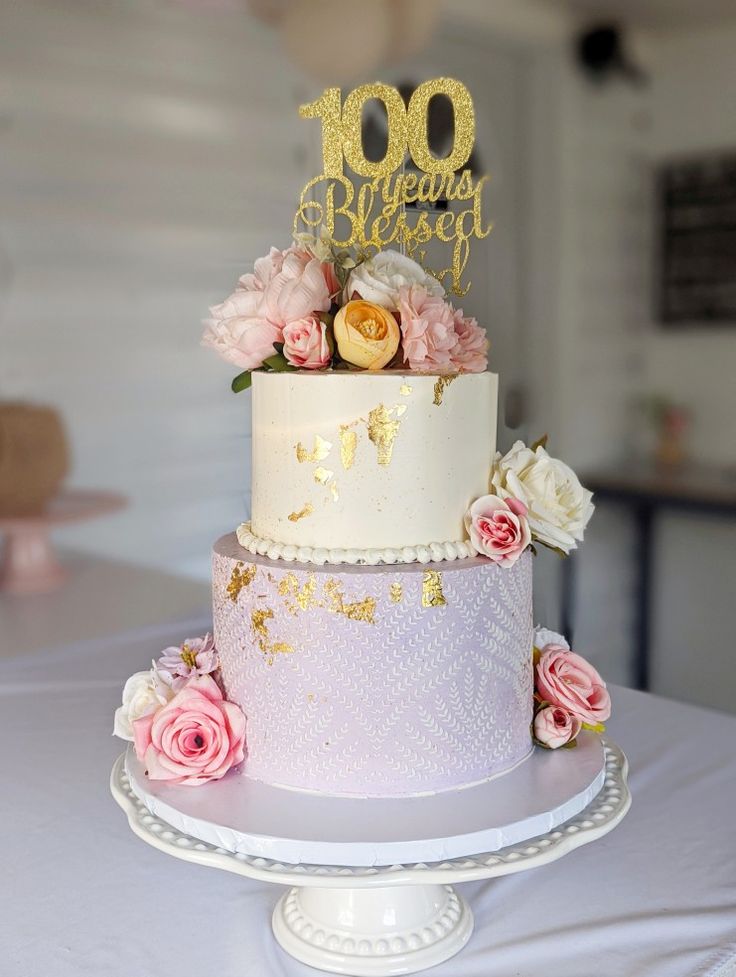 a three tiered cake with pink flowers and gold lettering on top is sitting on a table