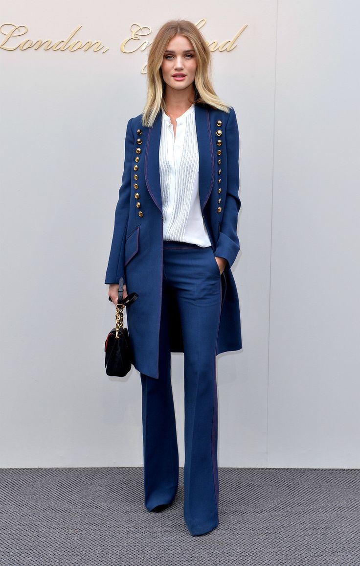 a woman standing in front of a white wall wearing blue pants and a blazer