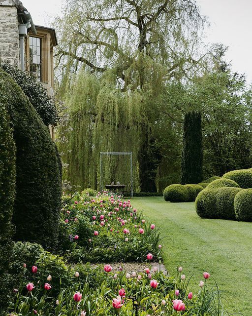 a lush green garden with pink flowers and trees in the background, next to a stone building