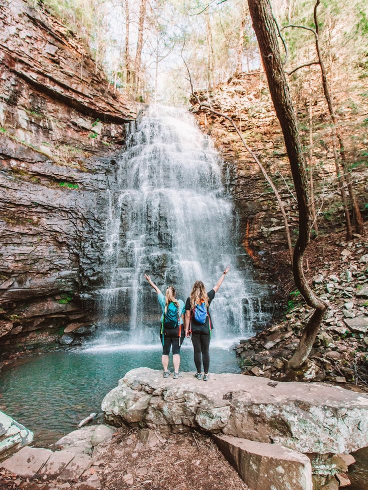 two women standing at the base of a waterfall with their arms up in the air