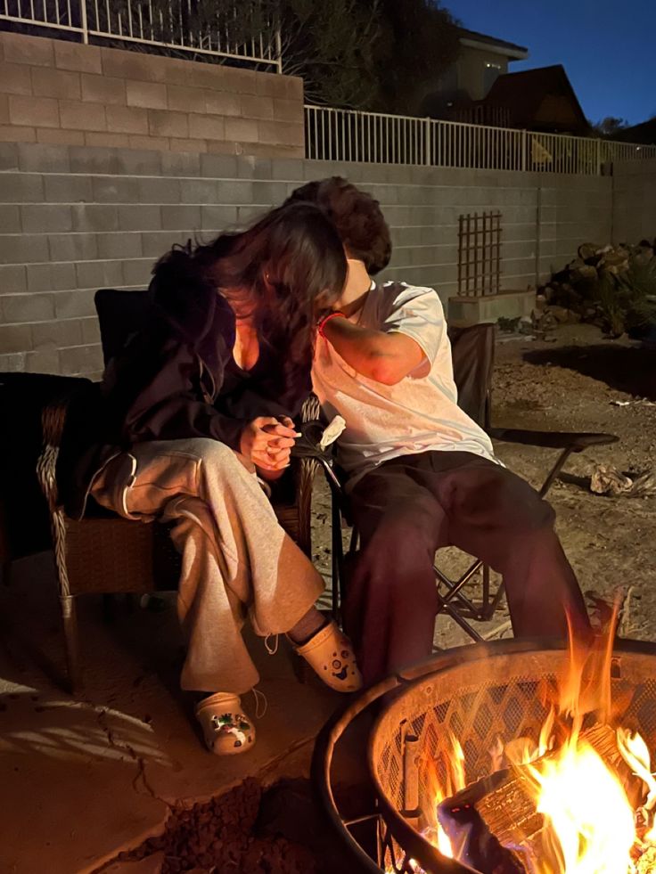 two people sitting next to each other in front of a fire pit with their faces close together