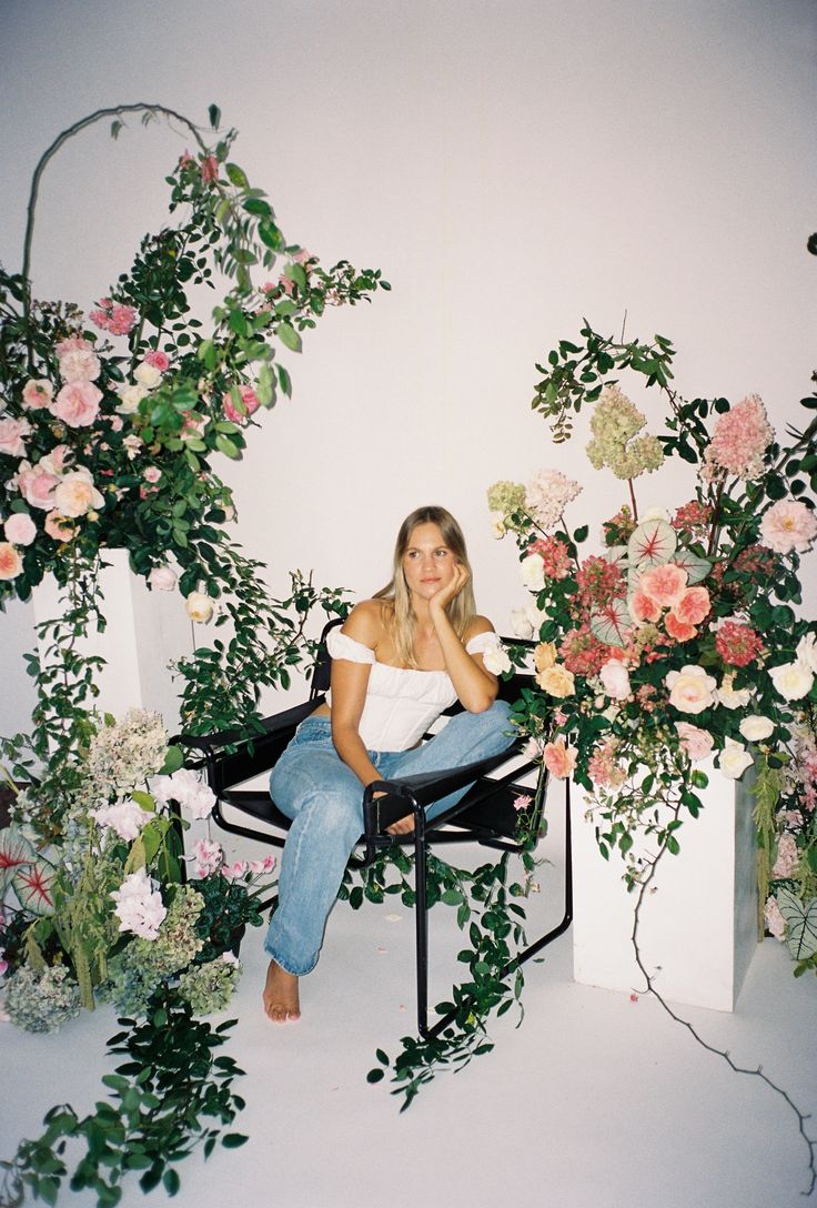 a woman sitting on a chair surrounded by flowers and greenery in front of a white wall