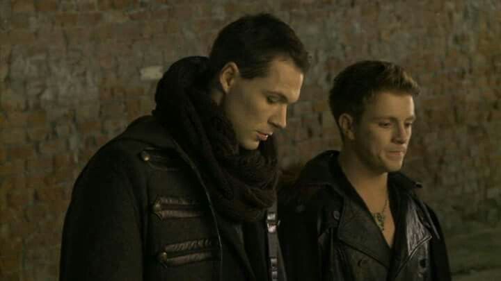 two men standing next to each other in front of a brick wall looking at their cell phones