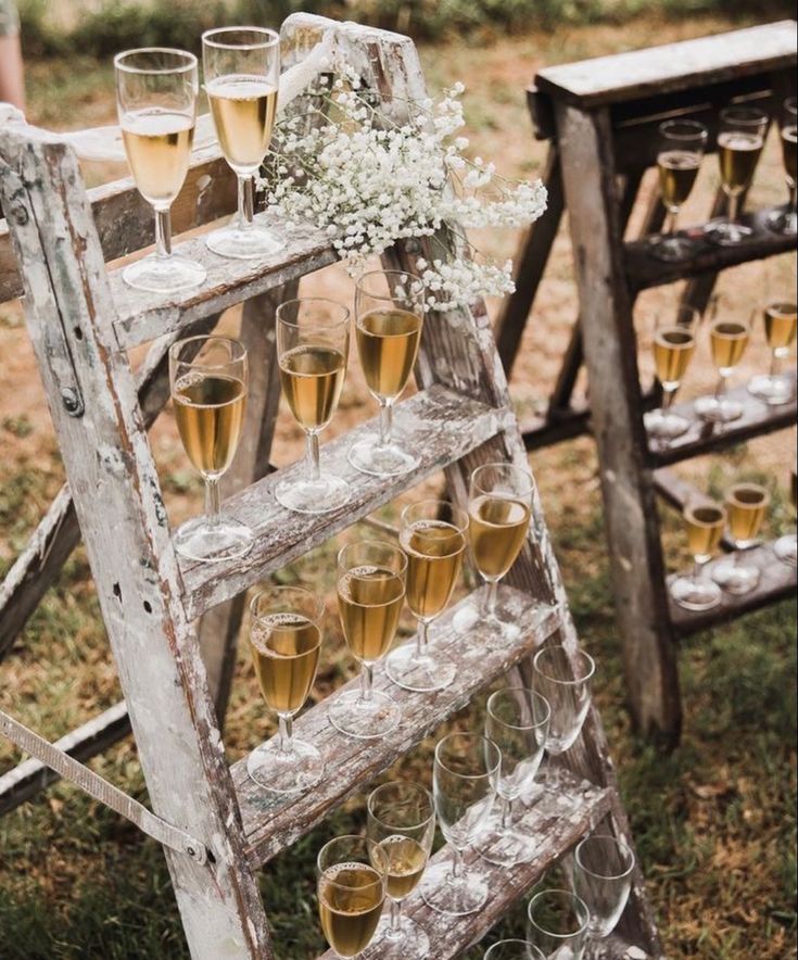 an old ladder filled with champagne glasses and flowers on top of it, sitting in the grass