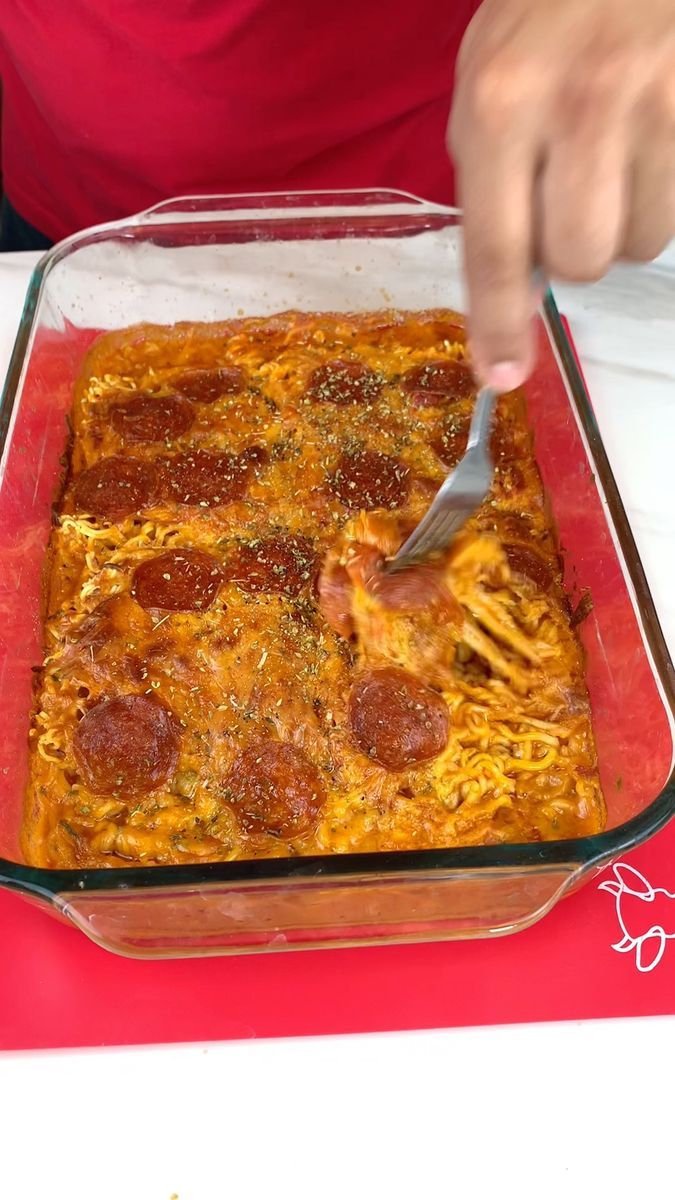 a person cutting into a pizza casserole in a glass dish on a table