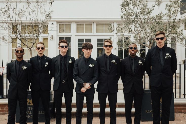 a group of men standing next to each other in front of a white building wearing black suits and sunglasses