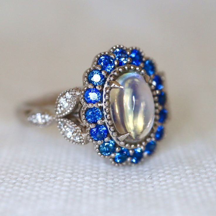 Mesmerizing Blue Moonstone Halo Ring. Moonstone engagement ring. Blue sapphires & diamonds. Edwardian Moonstone. 14K White Gold. US 7.25 A beautiful and unique moonstone ring, created with precision cut sapphires in 14K white gold, a luxurious and high-quality piece of jewelry, perfect to express your love. 💎 PREMIUM RING -- A brand new moonstone ring, personally designed and made with the highest quality of 14K white gold.  ✨ CLASSIC BRILLIANCE -- Designed with a beautiful blue moonstone at th Luxury Victorian Moonstone Ring Gift, Luxury Vintage Moonstone Ring As A Gift, Celestial Multi-stone Oval Moonstone Ring, Blue Birthstone Ring With Rose Cut Diamonds For Wedding, Blue Rose Cut Diamond Birthstone Ring For Wedding, Celestial Multi-stone Moonstone Oval Ring, Celestial Multi-stone Moonstone Ring, Blue Oval Multi-stone Cluster Ring, Blue Birthstone Ring With Rose Cut Diamonds For Anniversary