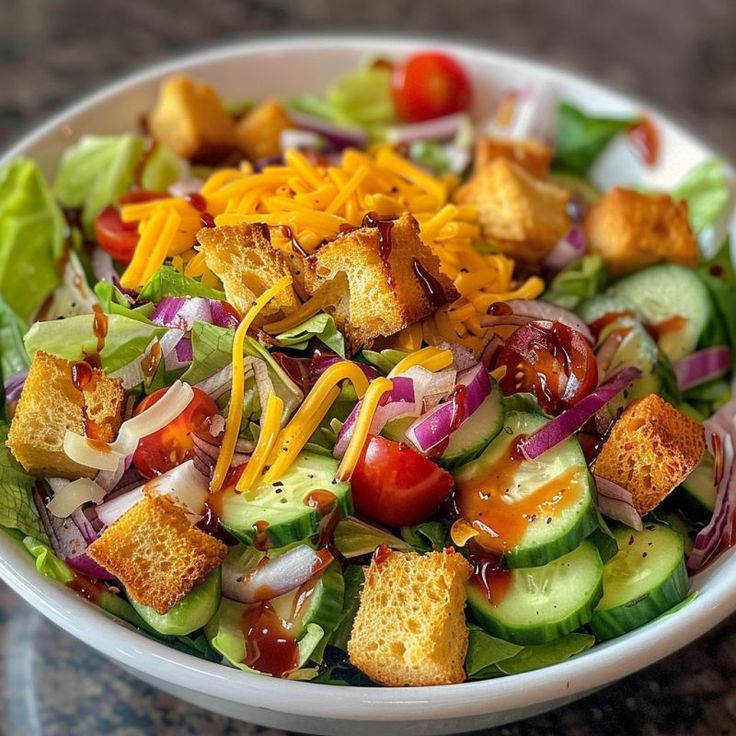 a salad with croutons, cucumbers, cheese and dressing in a white bowl