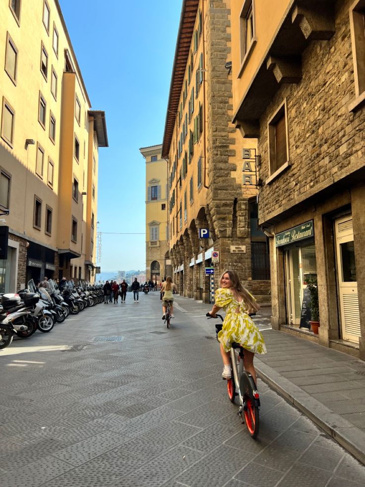 a woman riding a bike down a street next to tall buildings and parked motor scooters