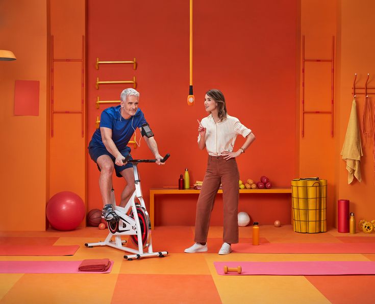 an older man and woman on exercise bikes in a brightly colored room with yoga mats