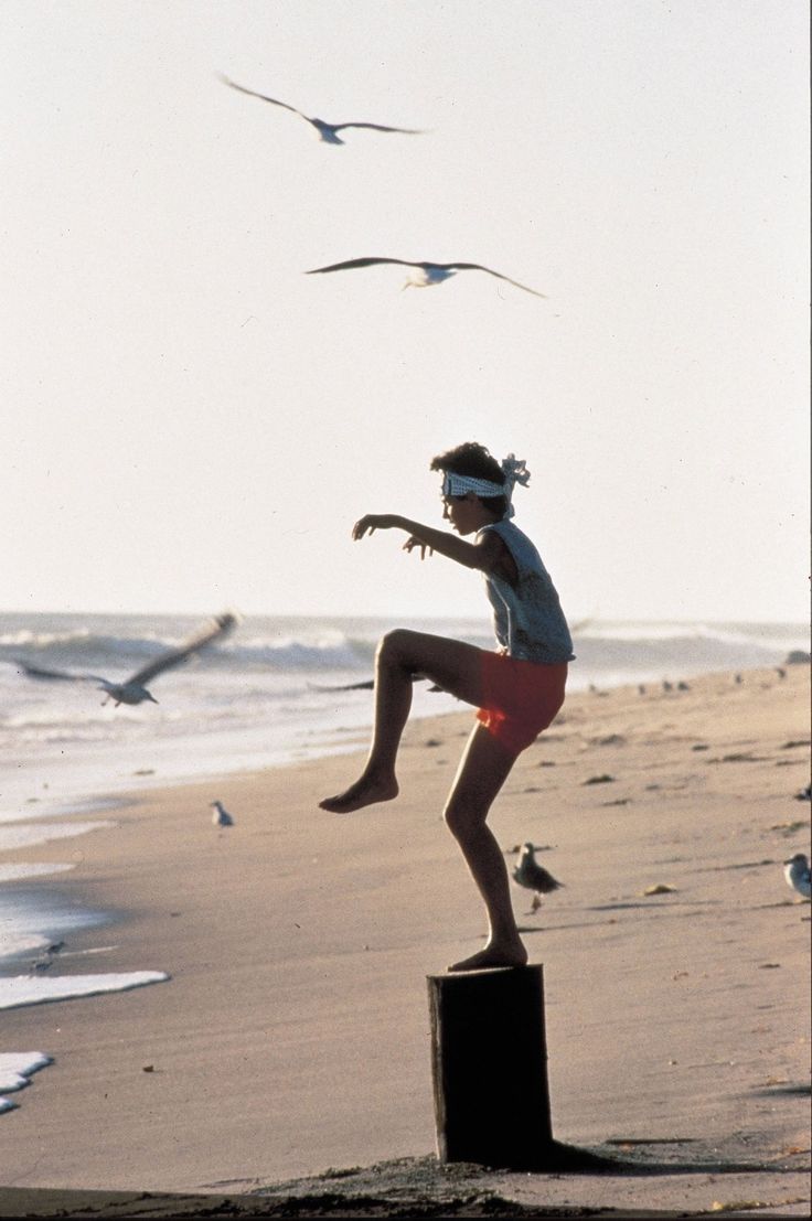 a woman standing on top of a wooden post near the ocean with seagulls flying around