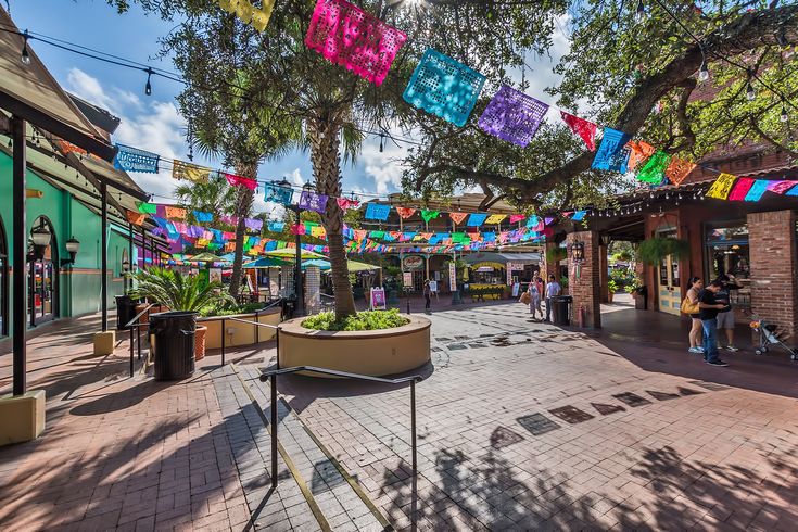 people are walking through an outdoor shopping area with colorful flags hanging from the trees above