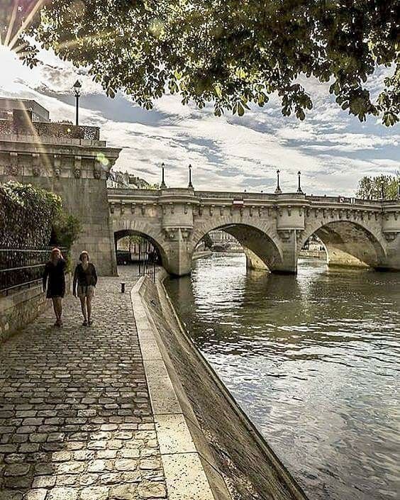 two people walking down a stone path next to a river with a bridge in the background