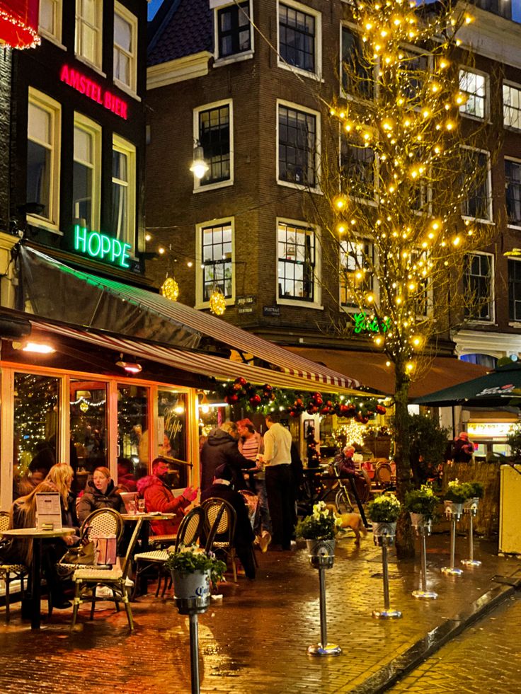 people are sitting at tables in front of a tree with christmas lights on the street