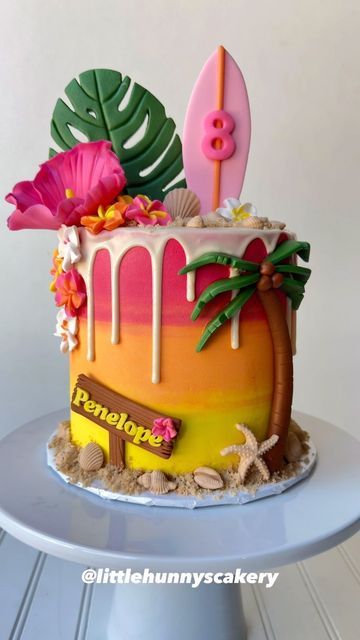 a colorful cake decorated with surfboards, flowers and palm leaves is on a table