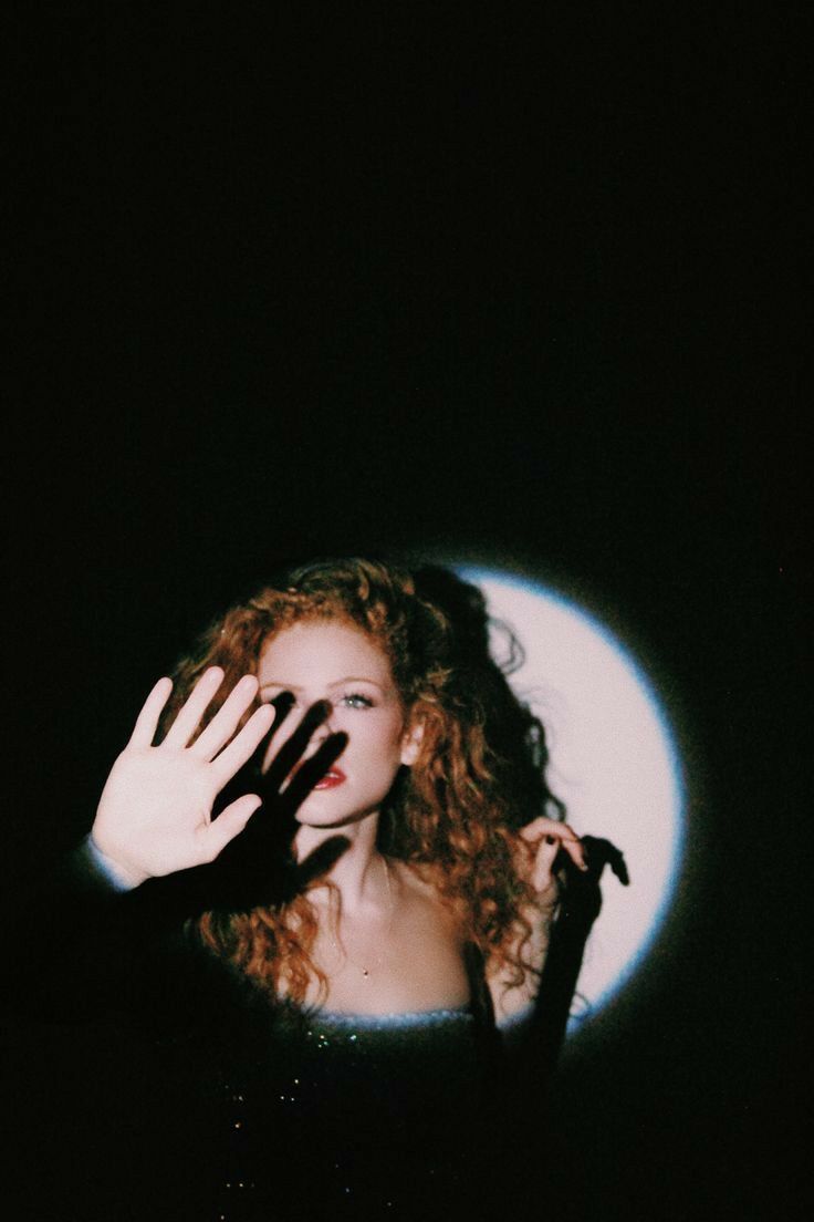 a woman holding her hand up to the camera in front of a dark background with light shining on her face