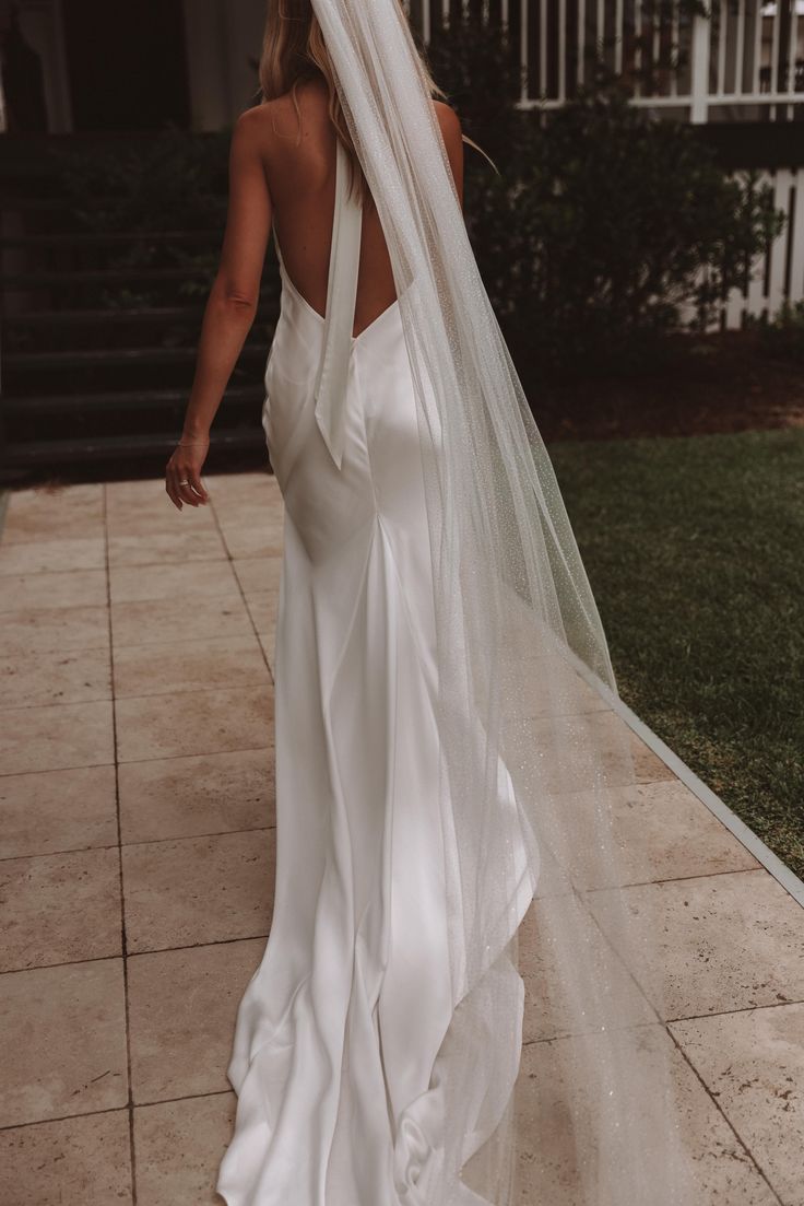 a woman in a white wedding dress and veil walking down the sidewalk with her back to the camera