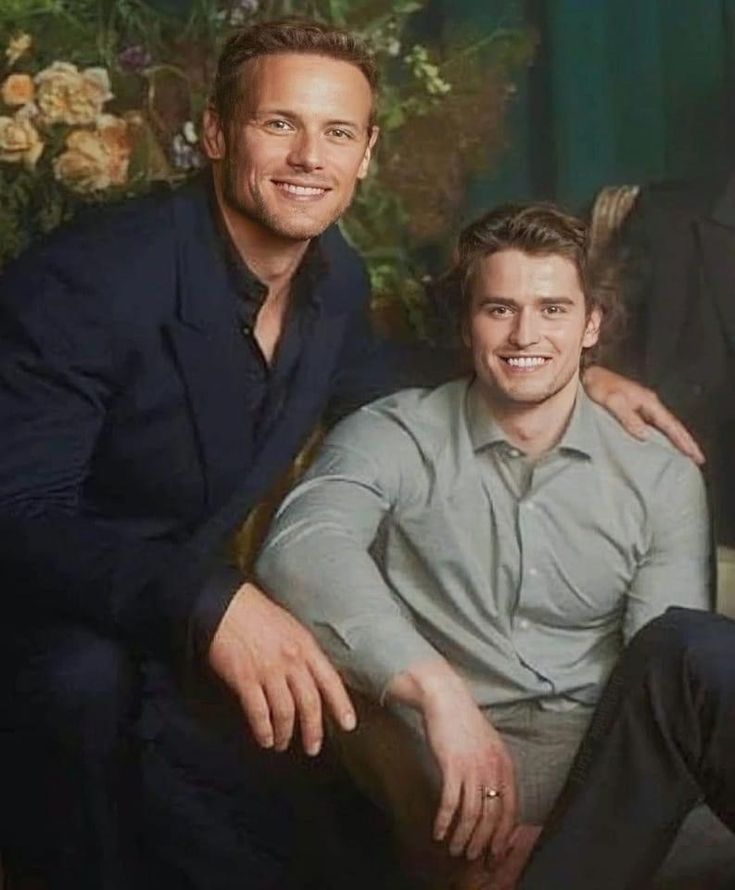 two men sitting next to each other in front of a floral wallpapered background