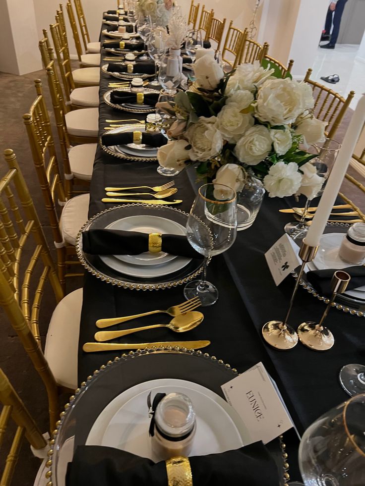 the table is set with black and gold place settings, silverware, and white flowers