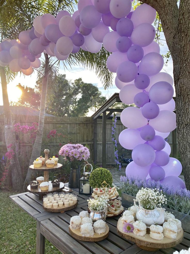 purple balloons are hanging over a table with cakes and cupcakes on it at a backyard party