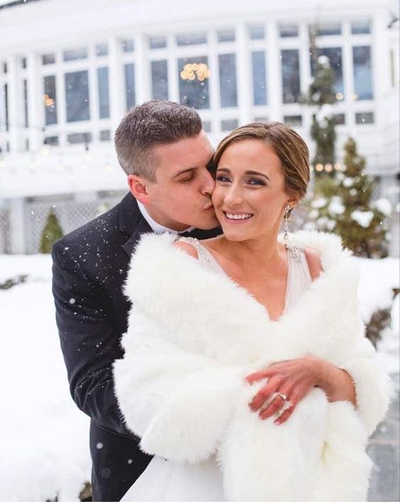 a bride and groom kissing in front of a white house covered in snow at their winter wedding