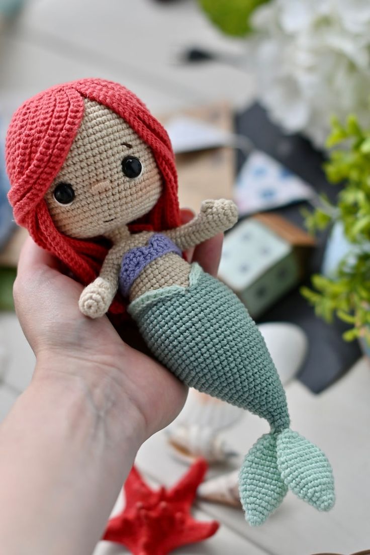 a hand holding a crocheted mermaid doll in front of a table with other items