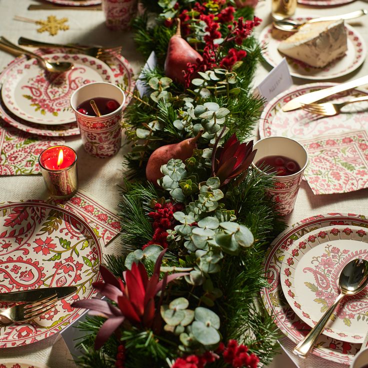 the table is set for christmas dinner with red flowers and greenery on it, along with silverware