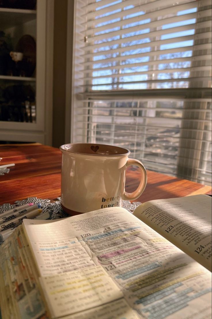 an open book and coffee cup on a table in front of a window with blinds