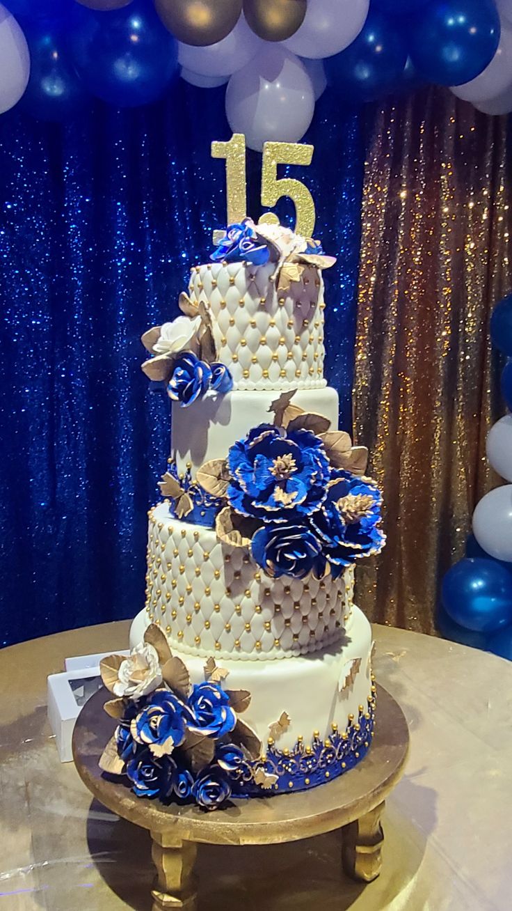 a wedding cake is decorated with blue flowers and gold accents on a table in front of balloons