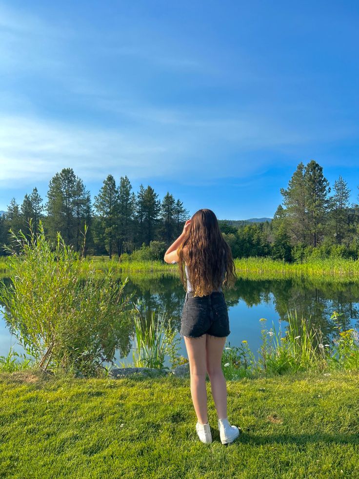 a woman standing on top of a lush green field next to a lake with trees in the background