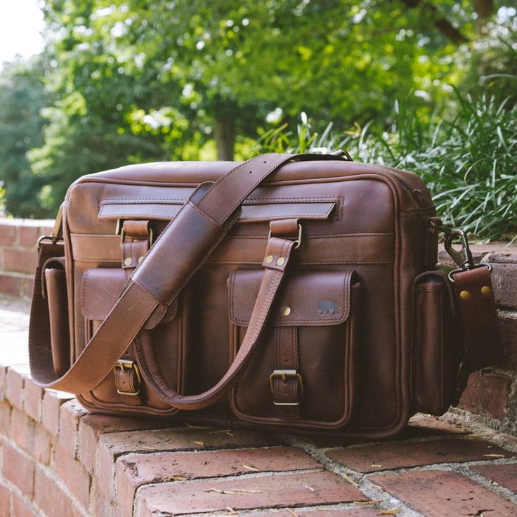 Our Roosevelt Leather Pilot Bag is crafted from water buffalo hide, with a green canvas lined interior, and genuine brass and iron hardware. It’s 16” long, 10” high, and 5” wide, the ideal size to hold a 15” laptop. Designed with four buckled exterior pockets and a roomy interior with zippered pockets, this bag is designed to securely hold the items you need to bring along on your travels. Built for comfort, this pilot bag has adjustable 2” wide shoulder straps, so you can comfortably carry it f Rectangular Travel Shoulder Bag With Brass Hardware, Rectangular Shoulder Bag With Brass Hardware For Travel, Brown Leather-lined Briefcase For Travel, Travel Satchel Briefcase With Brass Hardware, Leather Briefcase With Brass Hardware For Daily Use, Leather Shoulder Bag With Brass Hardware For Travel, Travel Satchel With Waxed Canvas Finish, Brown Travel Camera Shoulder Bag, Travel Laptop Bag With Leather Lining