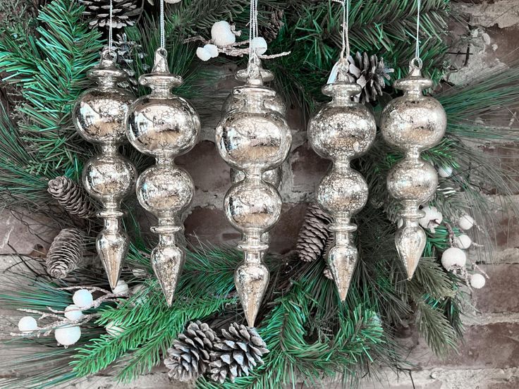 silver ornaments hanging from a christmas tree in front of a brick wall with pine cones