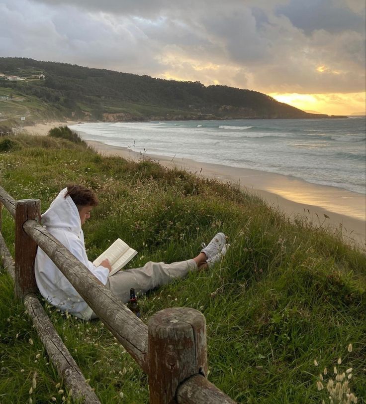 a person sitting on a bench reading a book by the beach at sunset or sunrise