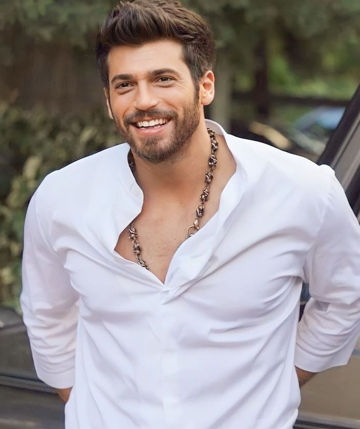 a man in white shirt and necklace standing next to a car smiling at the camera