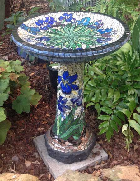 a bird bath in the garden with blue flowers on it's side and green leaves around it