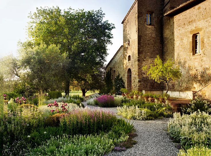 a stone building surrounded by lots of plants and flowers in the foreground is a gravel path leading up to it