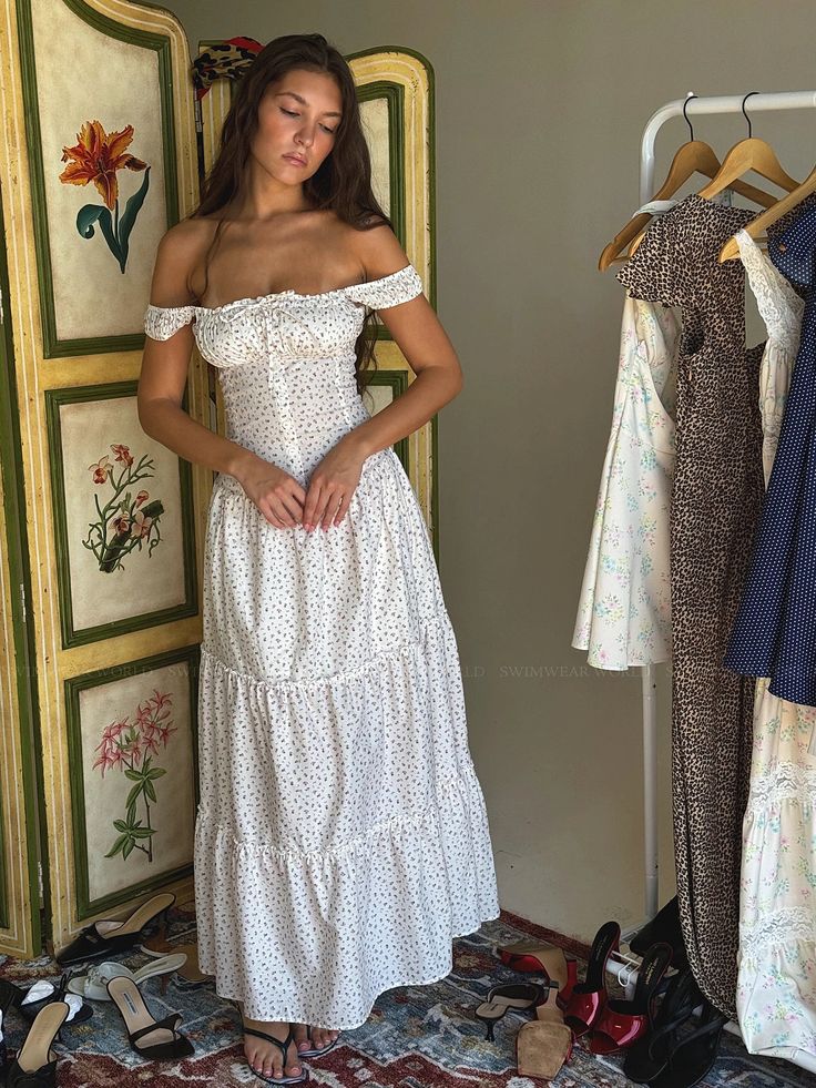 a woman in a white dress standing next to some clothes on hangers and looking at the camera