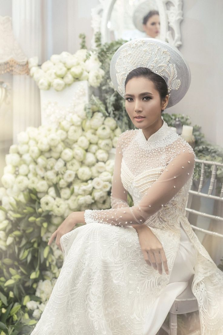 a woman in a white dress and hat sitting on a bench with flowers behind her