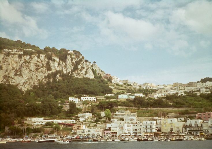 boats are docked in the water next to a hill with houses on it and buildings near by