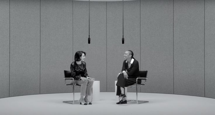 two people sitting in chairs facing each other on the same side of an empty room