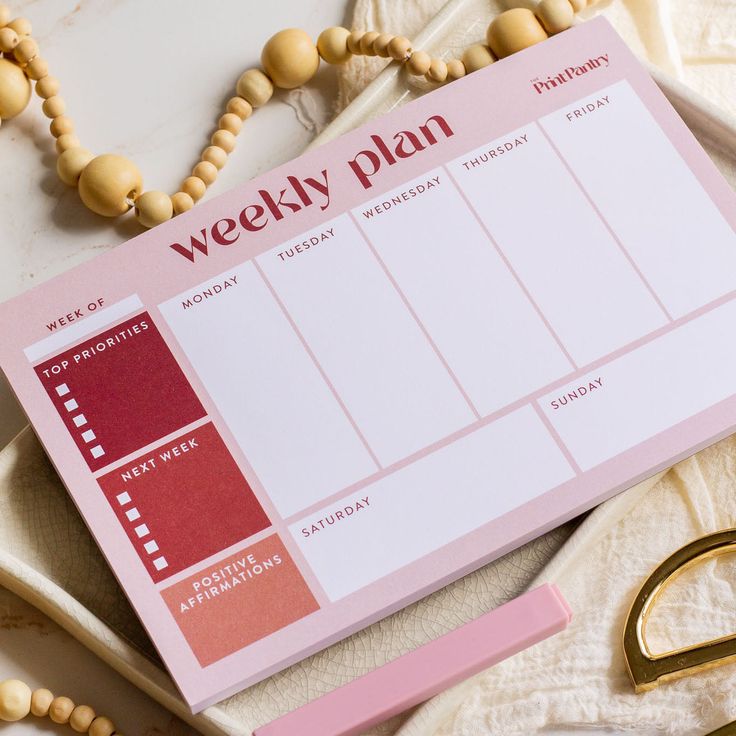 a pink weekly planner with gold scissors and beads next to it on a white surface