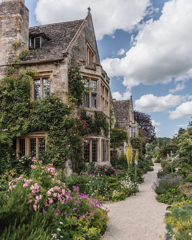 an old stone house surrounded by flowers and greenery on a sunny day with clouds in the sky