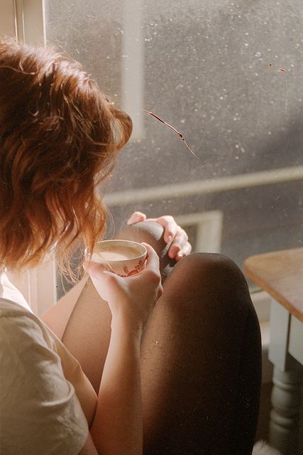 a woman sitting in front of a window drinking from a cup and looking out the window