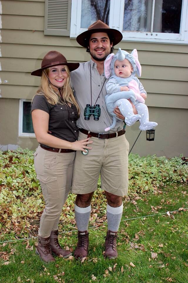 a man and woman standing in front of a house holding a baby wearing a hat