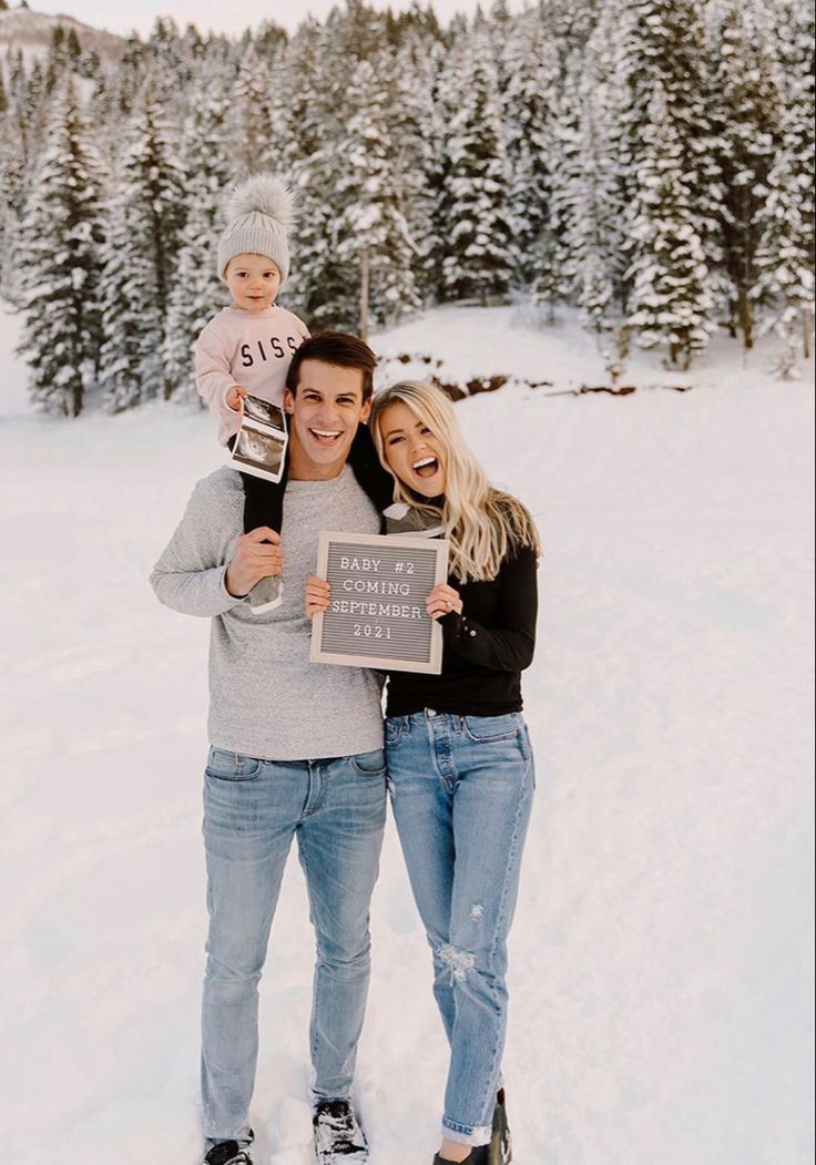 a man and woman pose for a photo while holding a sign in the snow with trees behind them