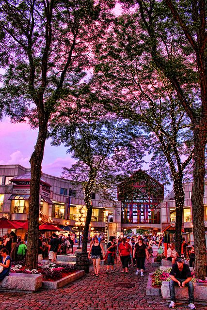 people are sitting on benches and walking around in the city at dusk, with trees lining the street