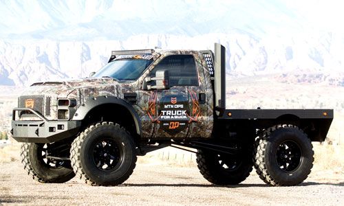 a camouflaged truck driving down a dirt road in front of mountains and desert grass