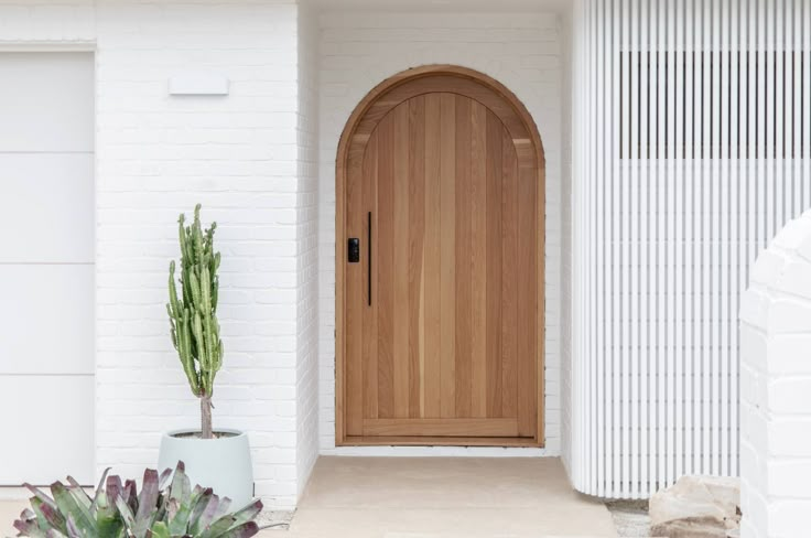 a house with a wooden door and cactus in the foreground, next to a white brick wall