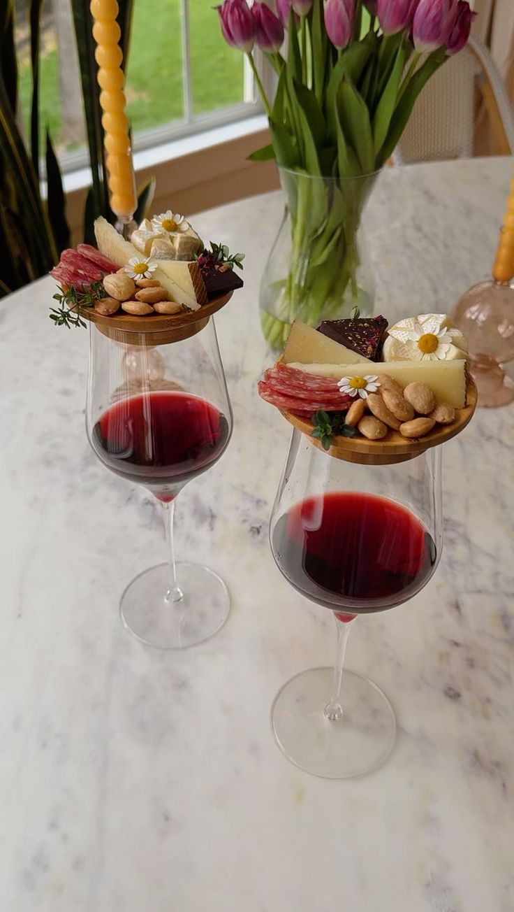 two wine goblets filled with red wine and cheese on a table next to tulips