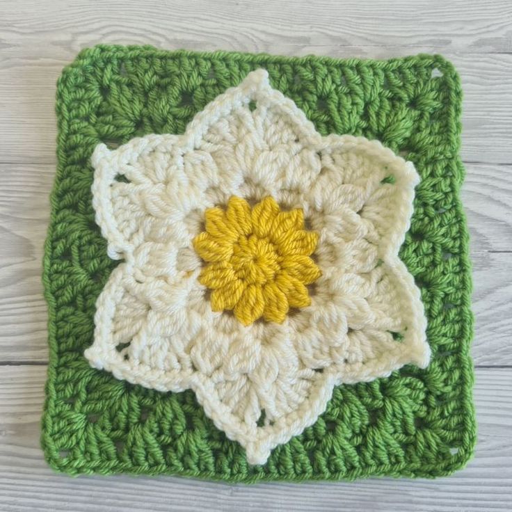 a crocheted square with a yellow flower on the center is sitting on a white wooden surface